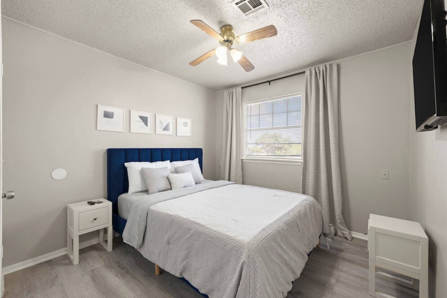 A bedroom with a bed, a nightstand, a ceiling fan, a window with curtains, and framed pictures on the wall.