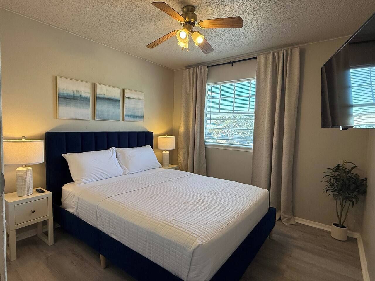 A bedroom with a bed, nightstands, a ceiling fan, a window with curtains, and a wall-mounted TV.