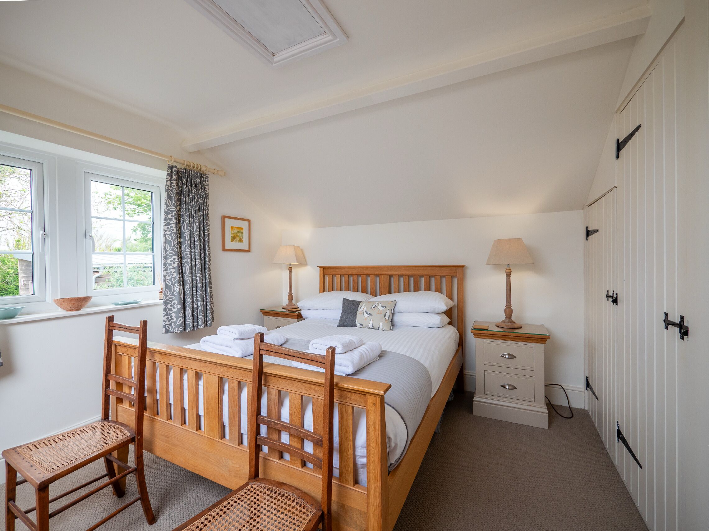 Une chambre à coucher avec un lit en bois, deux tables de chevet avec des lampes, une chaise, une fenêtre avec des rideaux et une armoire.