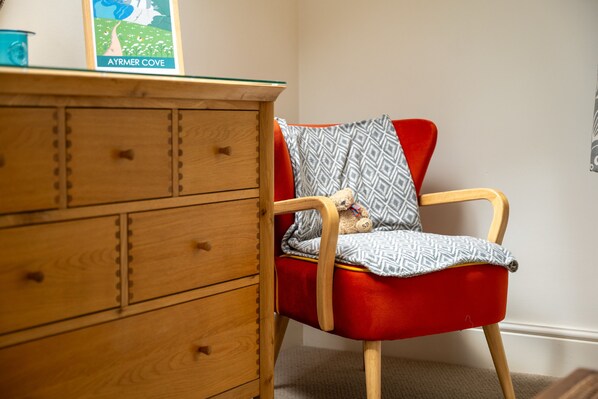 A wooden dresser with drawers, a red chair with a patterned cushion, and a framed picture titled 'AYRMER COVE' on top of the dresser.