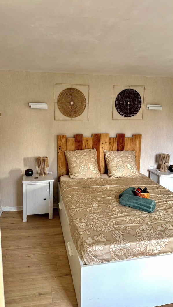 Ein Schlafzimmer mit Holz-Kopfteil, einer blumigen Tagesdecke, zwei Wanddekorationen, einem Nachttisch mit Lampe und einem Beistelltisch mit dekorativer Schale.