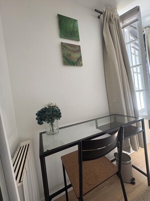 Une pièce comprenant un bureau avec un plateau en verre, une chaise noire, un radiateur, une fenêtre avec des rideaux et deux œuvres d’art accrochées au mur.
