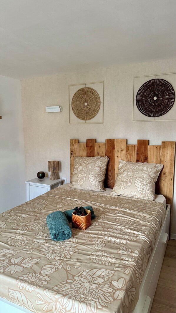 Un lit avec une literie à motifs floraux, une tête de lit en bois, deux œuvres d’art murales, une table de chevet avec une lampe et un panier décoratif.