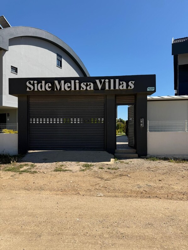 Un bâtiment moderne doté d’un grand portail de garage sur lequel on peut lire : « Entrée latérale, Melissa Villas ».