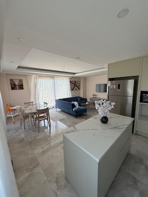 A modern kitchen with a built-in refrigerator, a dining area with a marble table and chairs, a blue sofa, and a large window with sheer curtains.