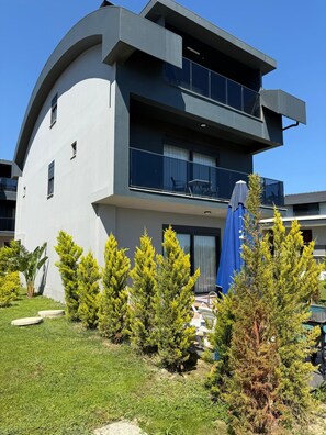 Un bâtiment moderne à deux étages, doté d’une façade incurvée, de balcons et d’un petit espace extérieur aménagé avec des sièges et un parasol bleu.