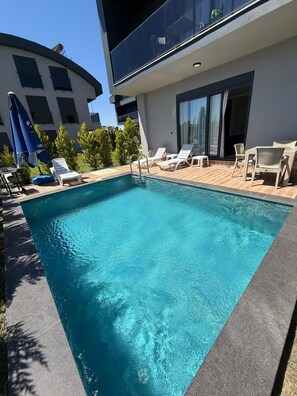 Ein rechteckiger Swimmingpool mit einer Holzterrasse, umgeben von Liegestühlen und Sonnenschirmen.