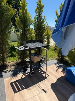 Un barbecue portable doté d’une grille en métal, d’un thermomètre intégré et d’un petit compartiment de rangement, installé sur une terrasse avec un plancher en bois et entouré de verdure.