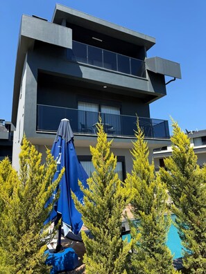 Ein modernes, zweistöckiges Gebäude mit Balkonen, einem Swimmingpool und umliegenden immergrünen Bäumen.
