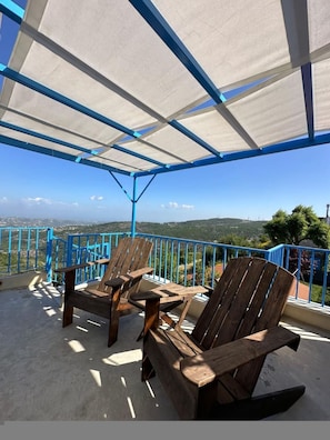 Un espacio de estar al aire libre con techo, barandillas azules, sillas de madera y vistas al paisaje.