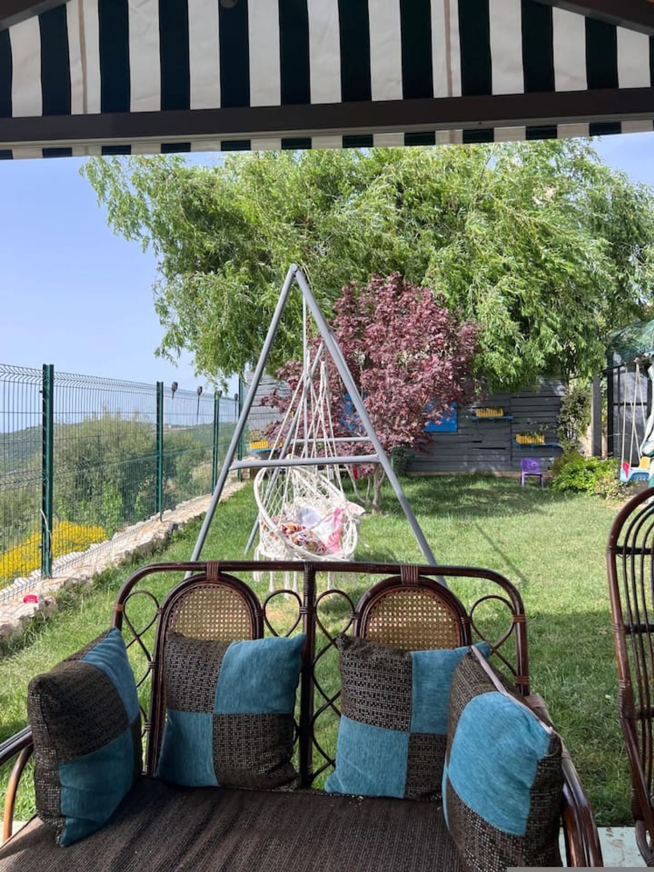 Un espacio exterior cubierto con asientos de mimbre y cojines, una mecedora y vistas a un jardín cercado.