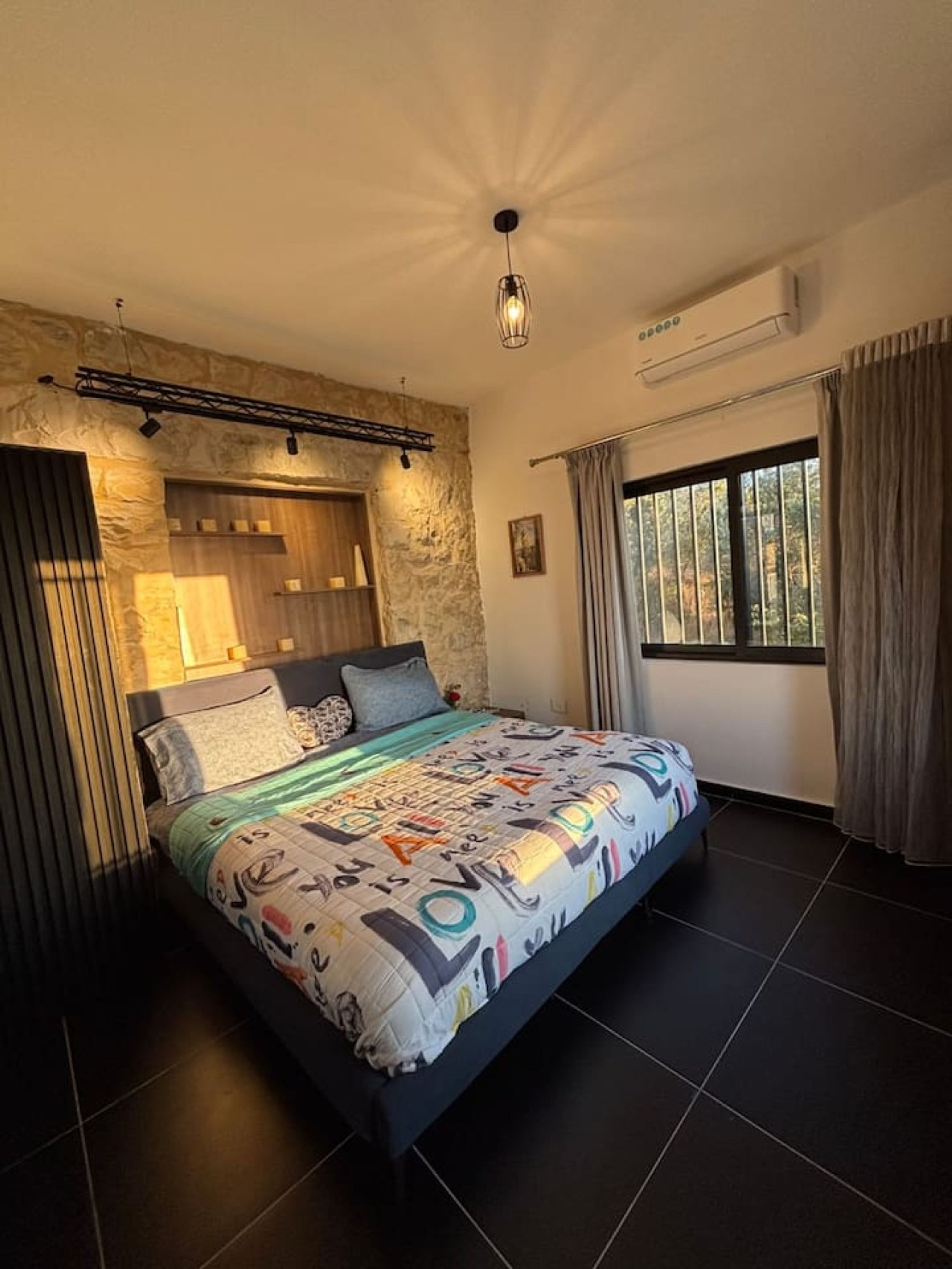 A bedroom with a bed, stone wall, wooden headboard, air conditioning, and a window with curtains.