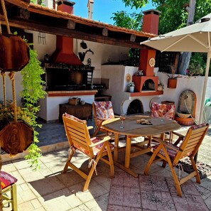 An outdoor patio with a round wooden table and chairs, a hanging plant, and a brick oven. - Traditional Stone House in a Peaceful Cretan Village - 20' from Heraklion Center (Epano Vatheia)