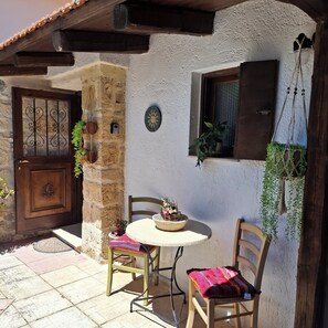 A stone building with a wooden door, a round table with chairs, and hanging plants. - Traditional Stone House in a Peaceful Cretan Village - 20' from Heraklion Center (Epano Vatheia)