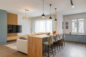 Ein modernes Wohnzimmer mit einem Holzesstisch, Barhocker, einem Flachbildfernseher und einem großen Fenster mit Vorhängen.