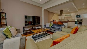 Una sala de estar moderna con un televisor de pantalla plana, sofás, una mesa de centro y una cocina al fondo.