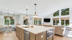 Eine geräumige Küche mit einer großen Kochinsel, Holzschränken und einem eingebauten Backofen. Es gibt Pendelleuchten, einen Essbereich mit Tisch und Stühlen sowie eine Wohnbereits mit einer Couch und einem Flachbildfernseher.