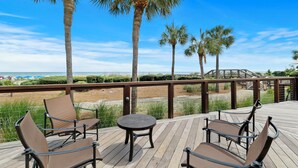 Eine Holzterrasse mit vier Stühlen und einem kleinen Tisch, mit Blick auf einen Strand mit Palmen und wolkenlosem, blauem Himmel.