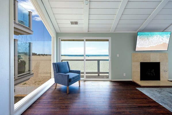 A room with a large window, a blue armchair, a fireplace, and a TV on the wall.