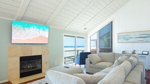 Un soggiorno con camino, una grande TV a schermo piatto, un divano beige e vista sull'oceano attraverso una porta scorrevole in vetro.