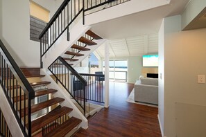 Un escalier avec des marches en bois et des rampes noires mène à un espace de vie comprenant un canapé blanc et offrant une vue sur l’extérieur.