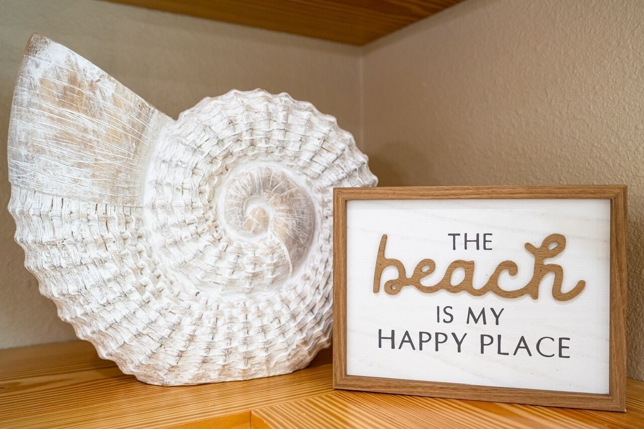 A framed sign with the text 'The beach is my happy place' next to a large white shell.