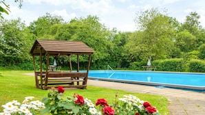 Un quiosco de madera, una piscina y macizos de flores con rosas y margaritas.