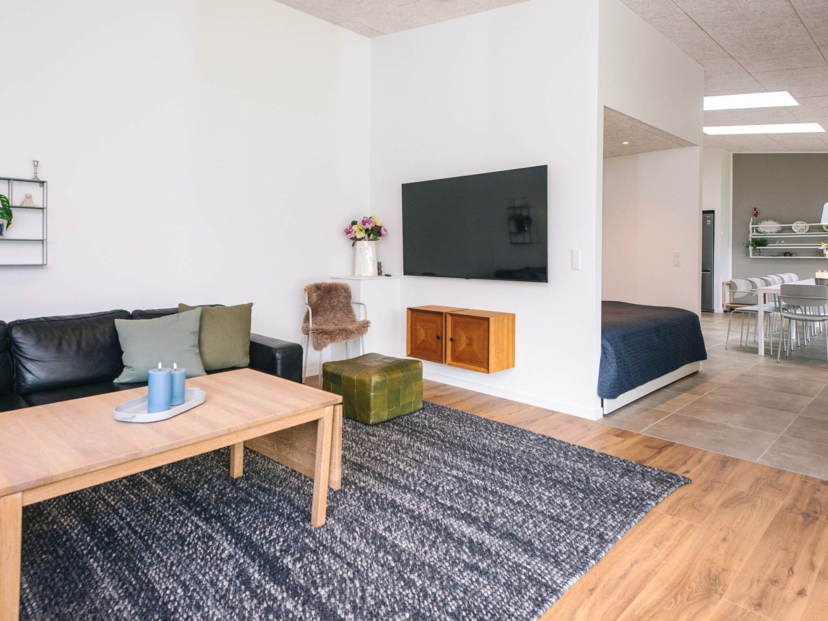 Een moderne woonkamer met een bank, een salontafel, een aan de muur bevestigde televisie, een klein kastje en een eethoek met stoelen en een tafel.