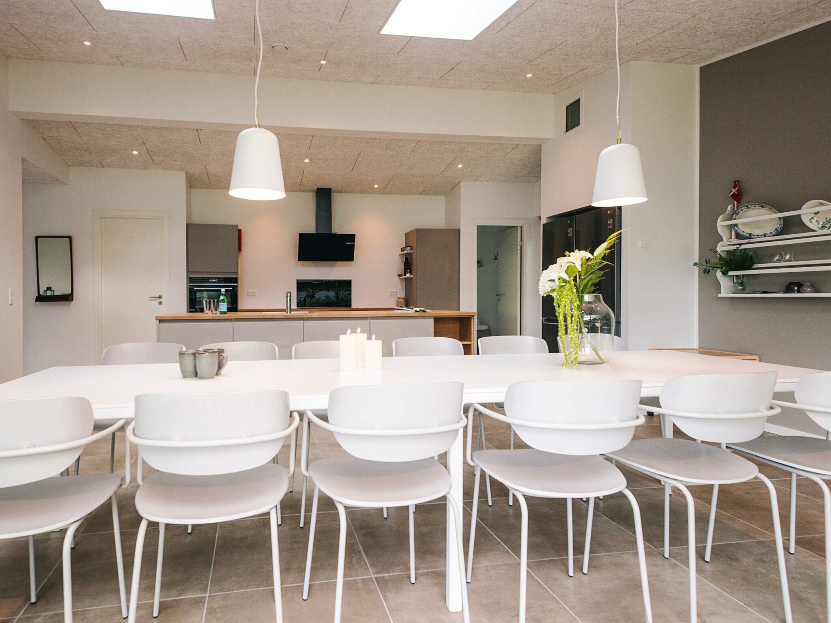 Een moderne eetruimte met witte stoelen en tafels, een keuken op de achtergrond en hanglampen.