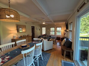 A spacious living area with a dining table, white chairs, a sofa, and a large window providing a view of the outdoors. - 4-bedroom Charming Cape Cod Cottage (Falmouth)