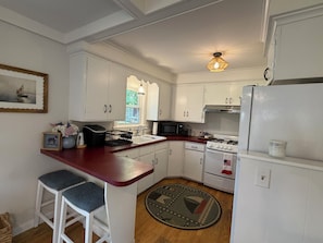A kitchen with white cabinets, a red countertop, a stove, a microwave, and a refrigerator. There are two bar stools and a rug on the floor. - 4-bedroom Charming Cape Cod Cottage (Falmouth)