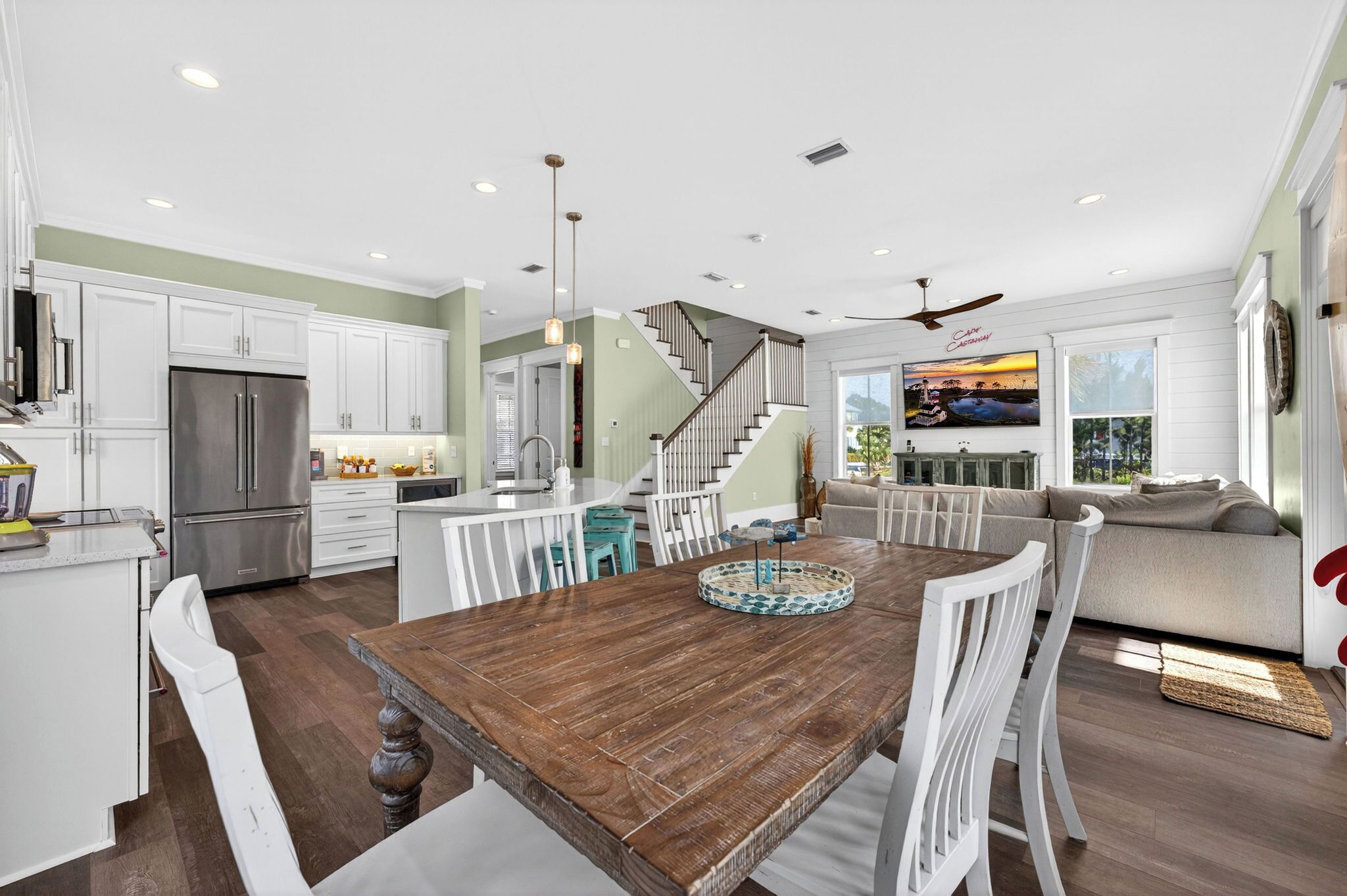 A modern kitchen with white cabinets, a stainless steel refrigerator, a wooden dining table with chairs, and a staircase leading to a living area with a sofa and a TV.