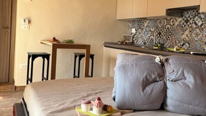 Une chambre à coucher comprenant un lit, des oreillers, une table de chevet, une petite table avec un plateau de nourriture, une chaise, un coin cuisine avec un mur recouvert de carreaux, et un plafond en briques.