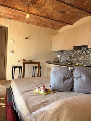 Ein Schlafzimmer mit Bett, Kissen, einem Nachttisch, einem kleinen Tisch mit Essensservit und einem Sessel, sowie einer Küchenzeile mit Fliesenrückwand und Ziegeldecke.