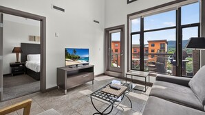 A modern living room with a TV, sofa, and coffee table, large windows with a city view, and a balcony with outdoor furniture. - 2 Bd Loft 603 @ Playa. Pool & Hot Tub (Kelowna)