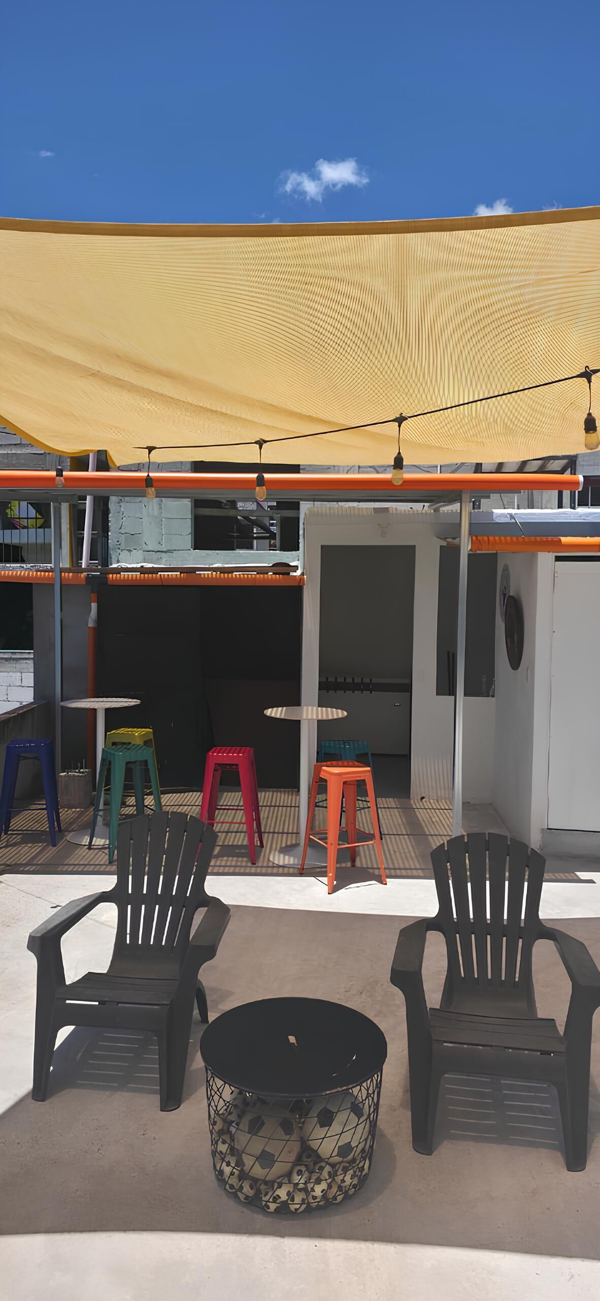 Un área de estar al aire libre con sillas y mesas, un toldo amarillo y un edificio al fondo.