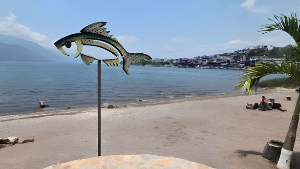 En strand med en skulptur, ett lugnt hav och en avlägsen stadsbild.