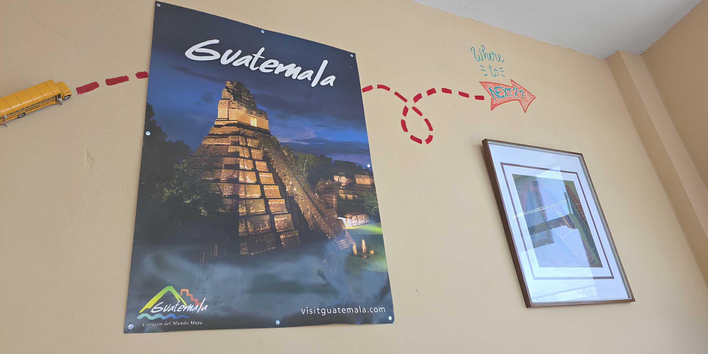 Paredes com pôster de Guatemala, uma moldura com fotografia e um ônibus de brinquedo amarelo.