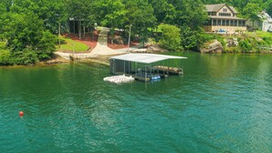 A lakeside dock with a covered structure, a boat, and a house in the background. - Lakecation on Smith Lake – Private boat launch! Private Dock! (Crane Hill)
