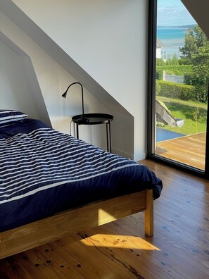 Ein Schlafzimmer mit einem Bett, einem kleinen Tisch und einem großen Fenster mit Blick auf eine grüne Landschaft und das Meer.