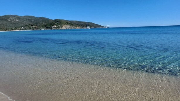 Uma praia com águas cristalinas, uma orla arenosa e uma paisagem montanhosa ao fundo.