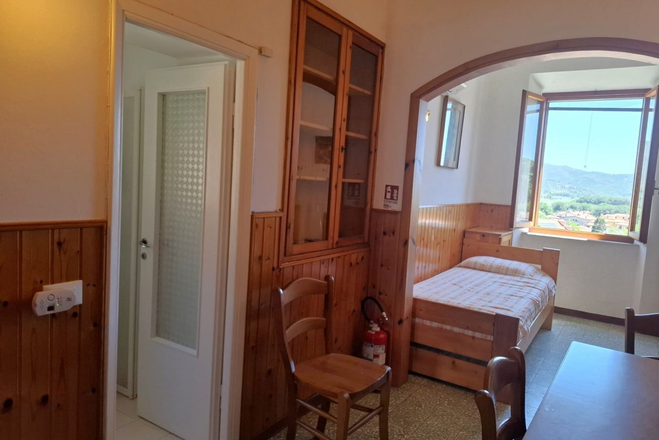 A bedroom with a wooden wardrobe, a bed, a chair, and a view of a landscape through a window.