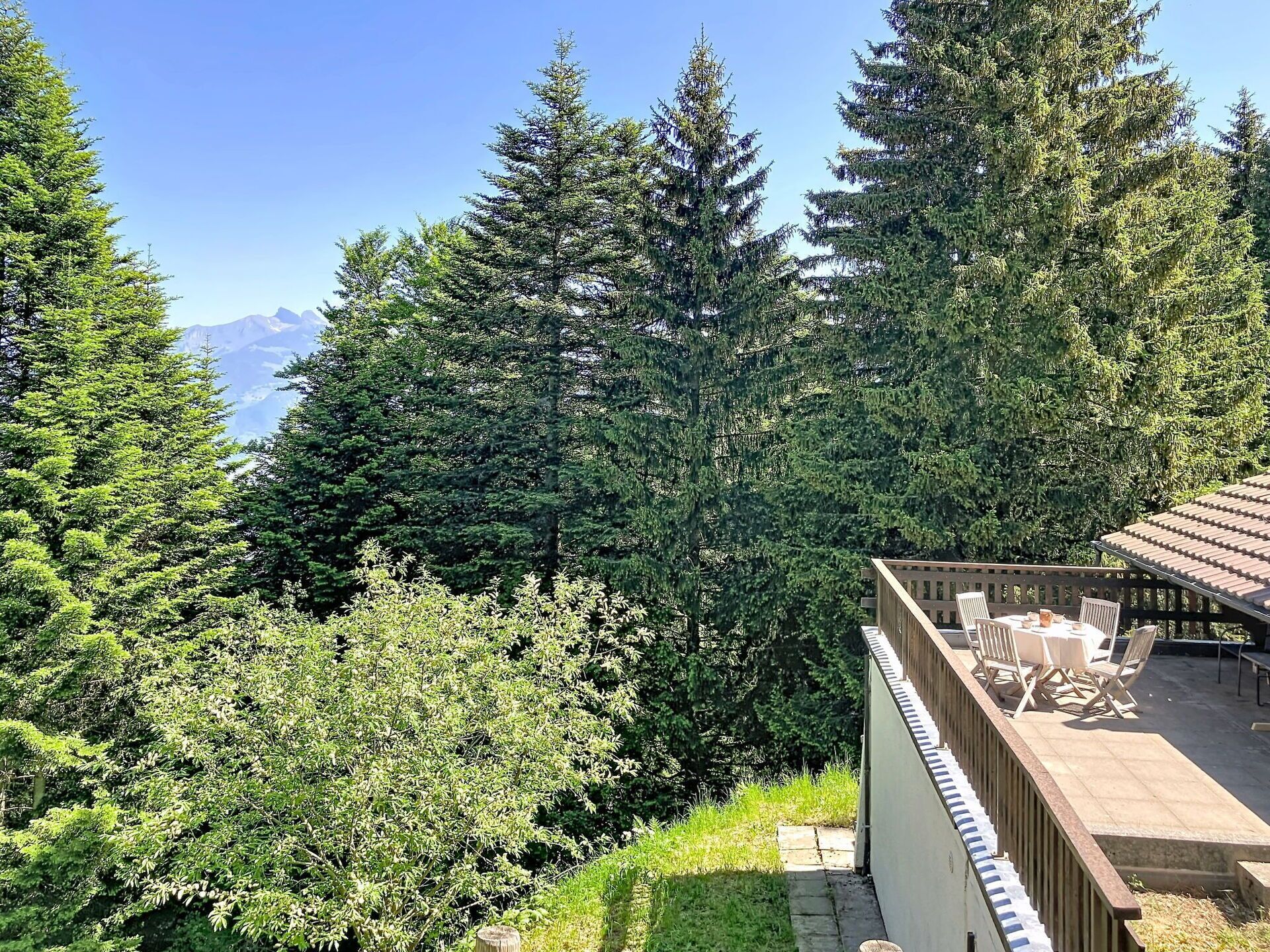 Blick auf eine gebirgige Landschaft mit dichtem Wald und einer Holzterrasse mit Gartenmöbeln.