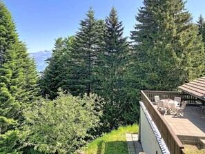 Blick auf eine gebirgige Landschaft mit dichtem Wald und einer Holzterrasse mit Gartenmöbeln.
