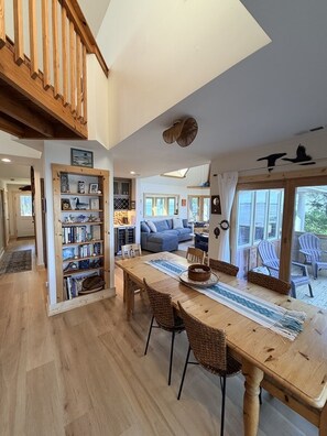 Un vaste espace de vie comprenant une table à manger en bois, des chaises en osier, une bibliothèque et une vue sur l’extérieur.