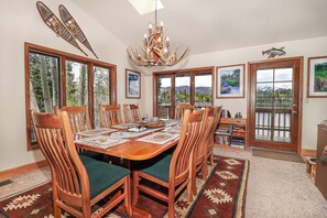 A dining room with a large wooden table, chairs, a chandelier, and large windows with mountain views.
