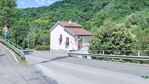 Ein kleines, weißes Gebäude mit rotem Dach, eine Straße mit Schutzrail und ein Hügel mit dichtem Grün im Hintergrund.