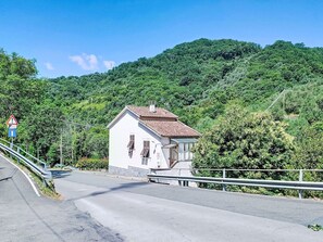 Un petit bâtiment blanc avec un toit rouge, une route bordée d’une glissière de sécurité et, au loin, une colline couverte d’un feuillage vert et dense.