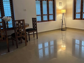A room with a dining table, chairs, a lamp, and large windows with blinds. - Villa Lemon Garden (Varkala)