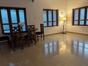 A room with a dining table, chairs, a floor lamp, and windows with wooden frames. - Villa Lemon Garden (Varkala)